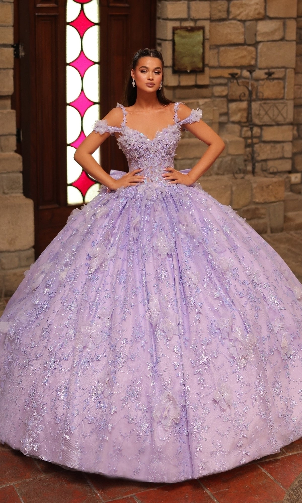 A woman poses indoors in front of a stone wall and wooden door wearing the Amarra 54243 off-shoulder quinceanera dress, featuring an elaborate lavender design with 3D floral appliques.